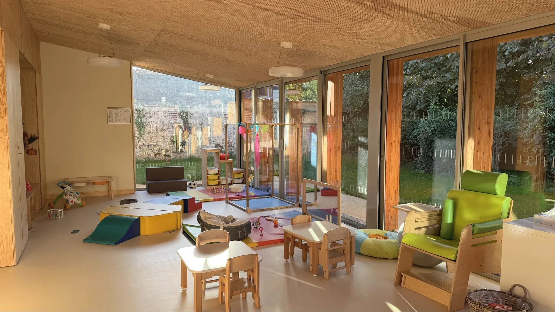 Salle de vie lumineuse de la crèche avec tables et petites chaises, jeux au sol, structures en bois et baies vitrées donnant sur le jardin.