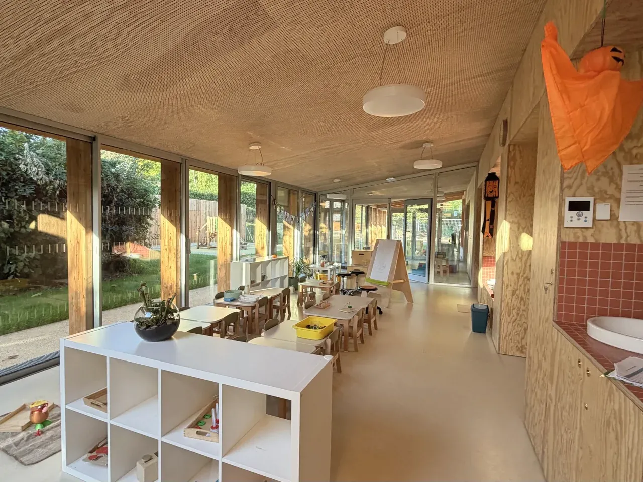 Espace d'activités avec petites tables, chaises en bois et étagères, le long de grandes fenêtres donnant sur le jardin.