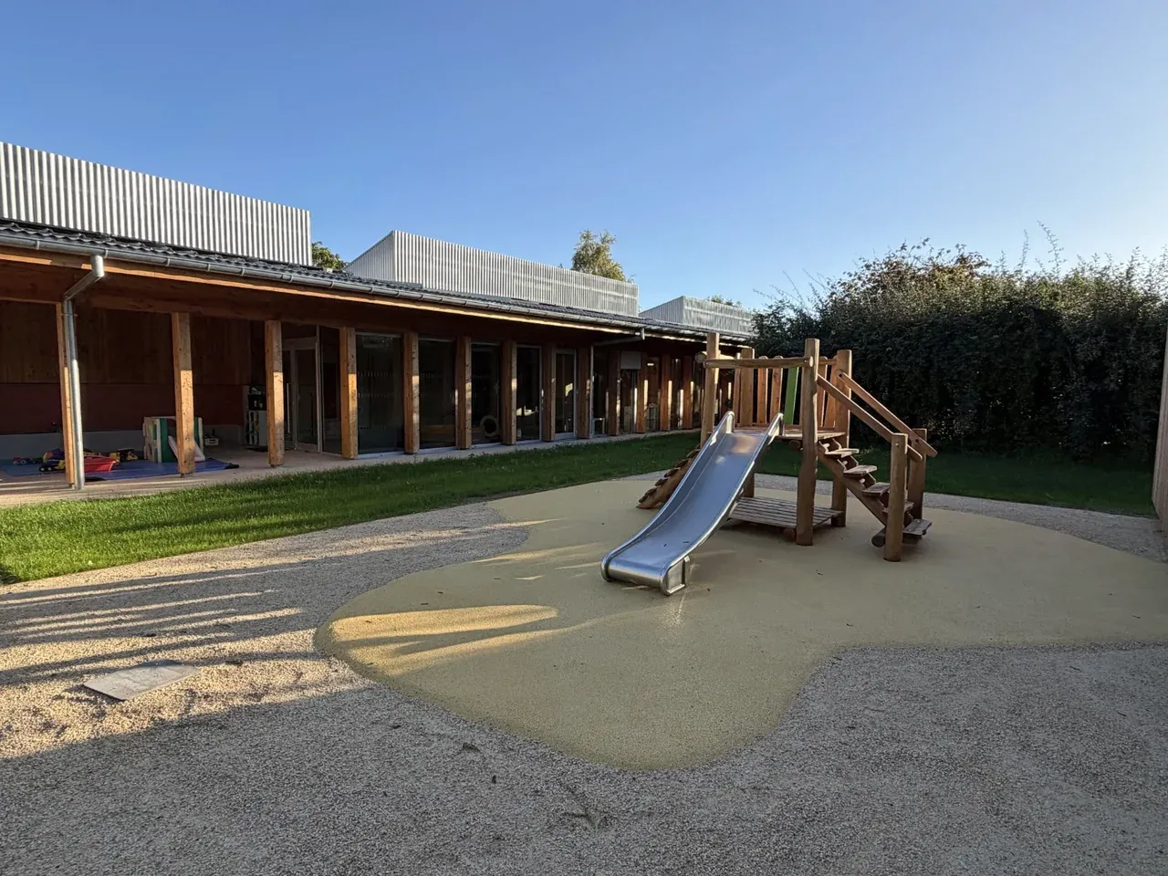Aire de jeux extérieure avec un petit toboggan en bois et des pelouses devant le bâtiment de la crèche.